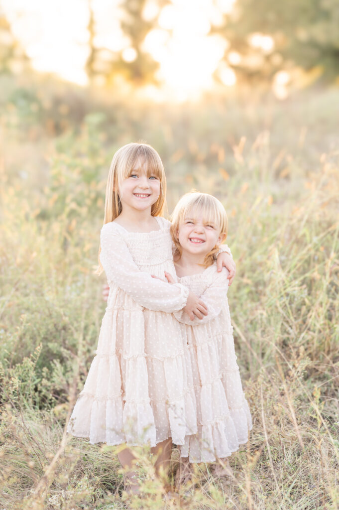 Two young sisters hugging each other in matching neutral dresses and smiling at the camera in a tall grass field with the golden sun setting behind them. 