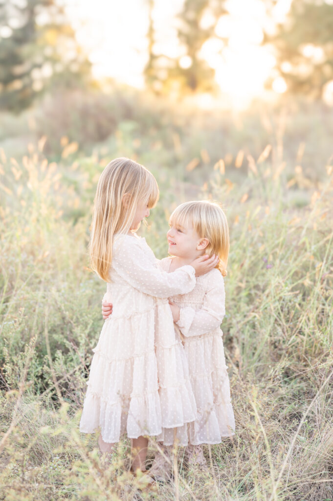 Two young sisters in matching dresses embracing in a field with a golden sunset behind them.