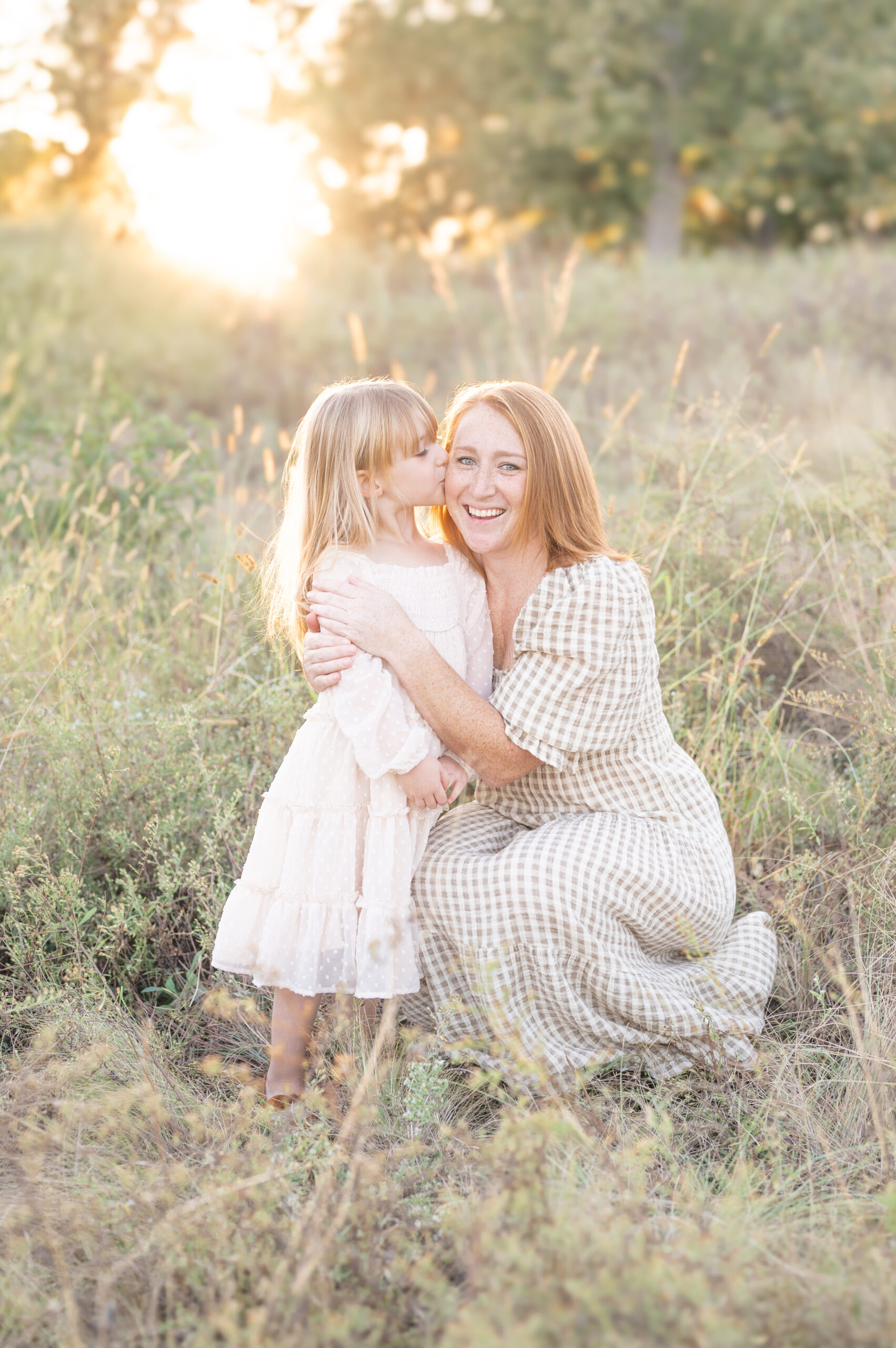 A mother kneeling down by her young daughter in the tall grass and her daughter kissing her cheek with the sun setting behind them. 