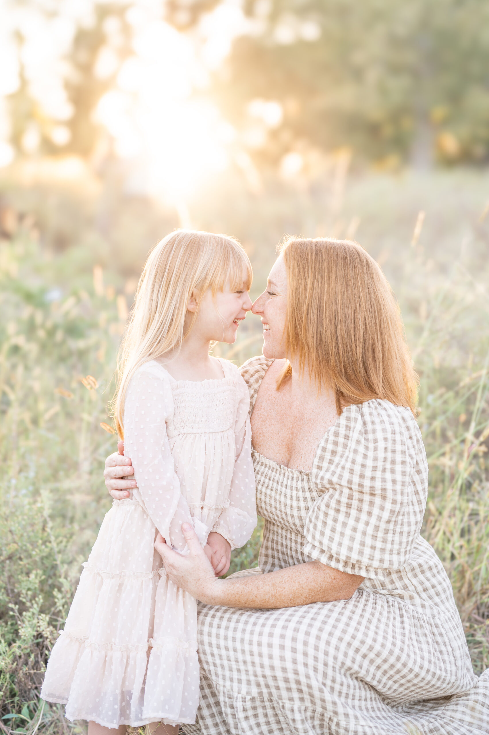 A mother in a maxi neutral dress kneels next to her young daughter in a cream dress touching noses and smiling with the golden sun setting behind them.