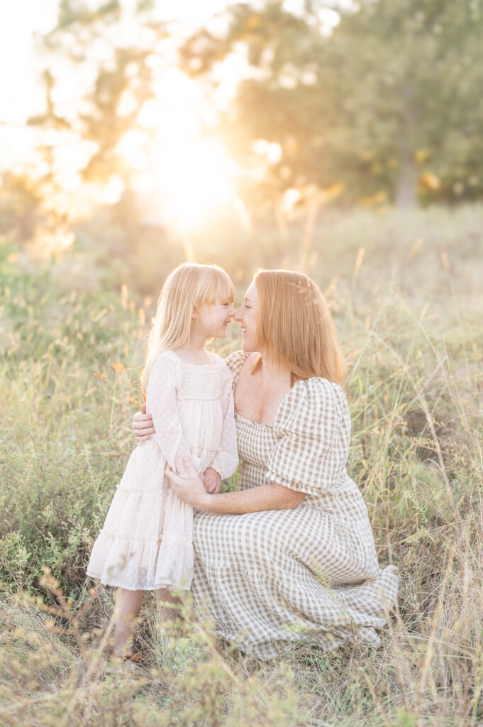 A mother kneeling down and touching noses with her young daughter in the tall grass with the sun setting behind them by a Raleigh Family Photographer. 