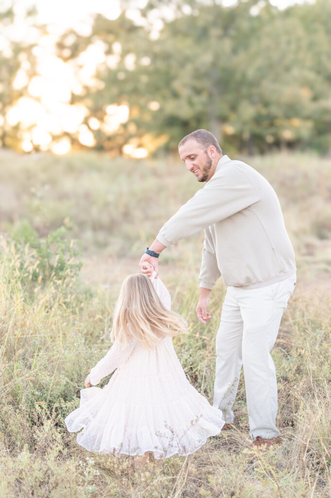 A father and his young daughter in a tall grass field. His daughter holds his hand and twirls around. 