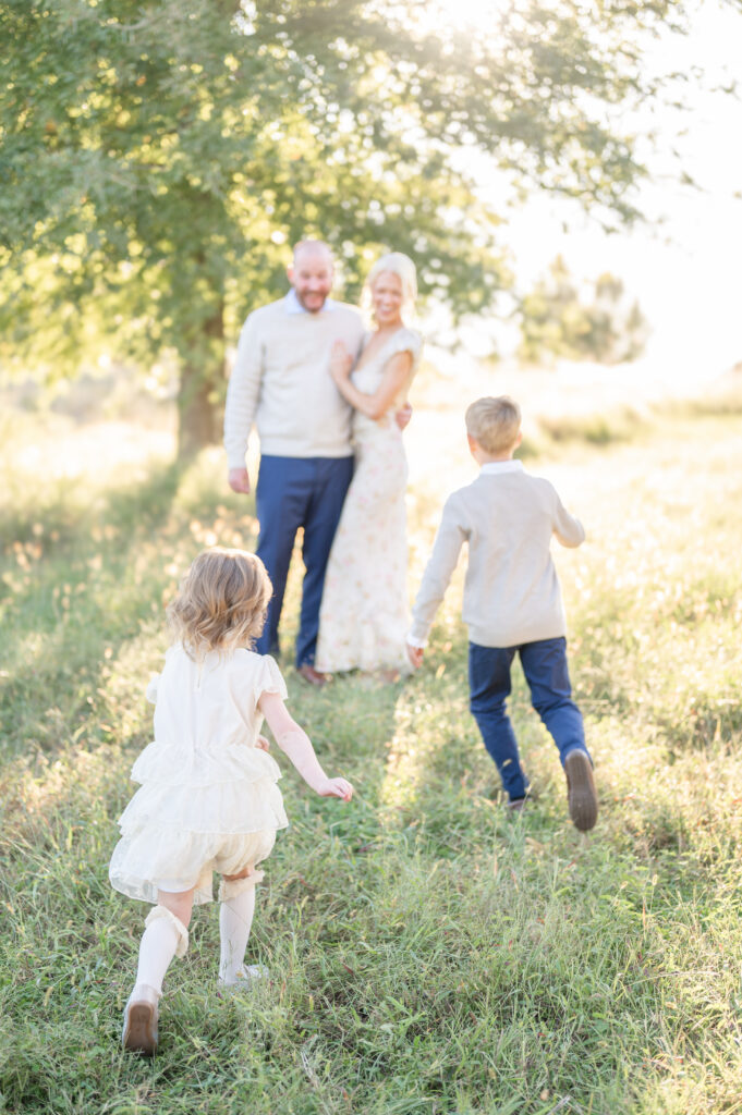 A mother and a father embracing with their backs to the sunset while their two young kids run towards them.