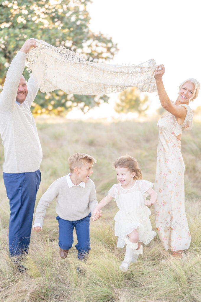 A mother and a father hold a blanket up in the air while smiling at the camera while their two young children run underneath holding hands and smiling at each other. 