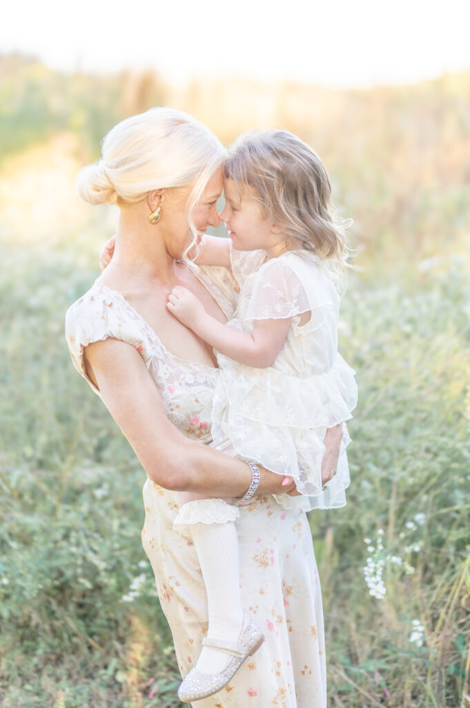 A mother holds her young daughter dressed in a white dress and knee high socks while resting their foreheads together and smiling at each other. 