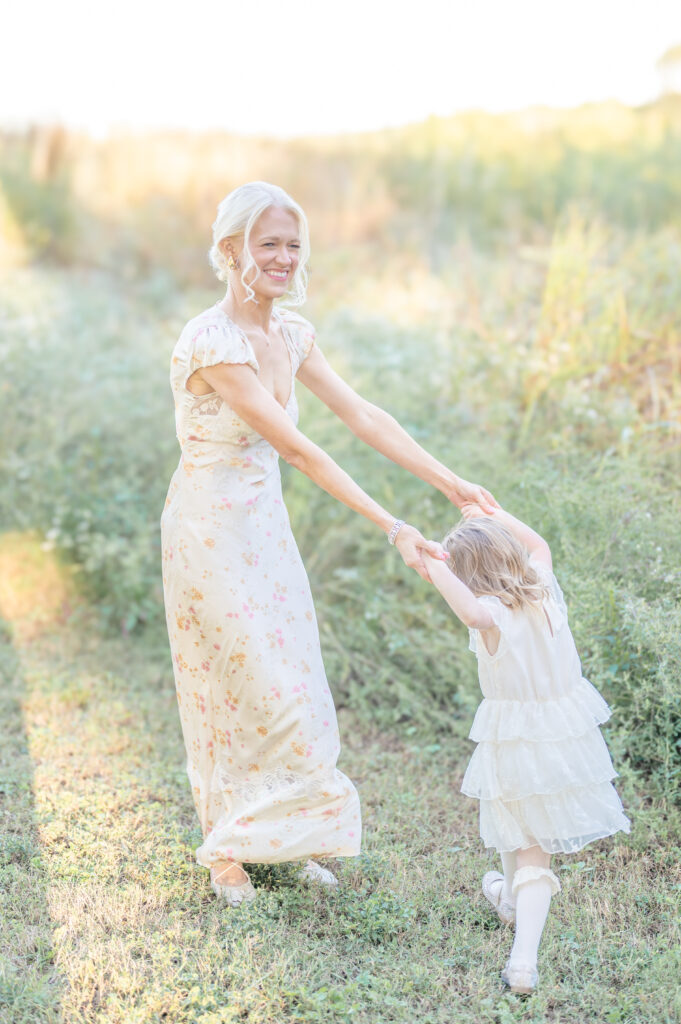 A mother in a light colored floral dress holds hands with her young daughter while spinning in a circle in a field at sunset.