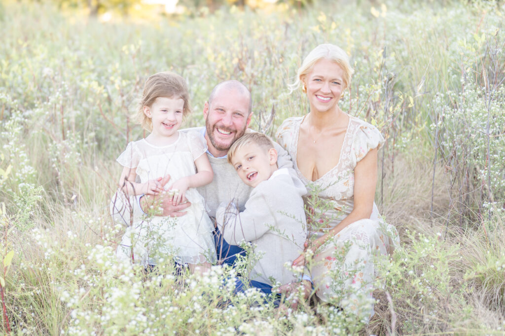 A portrait of a young family of four sitting in grass field at sunset smiling and enjoying each others company.